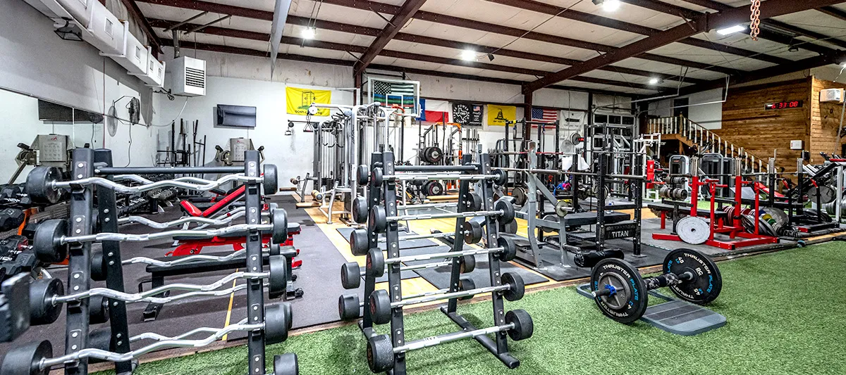 The lifting floor at Spicewood Fitness: racks, benches and loaded barbells across the turf under a high metal roof.